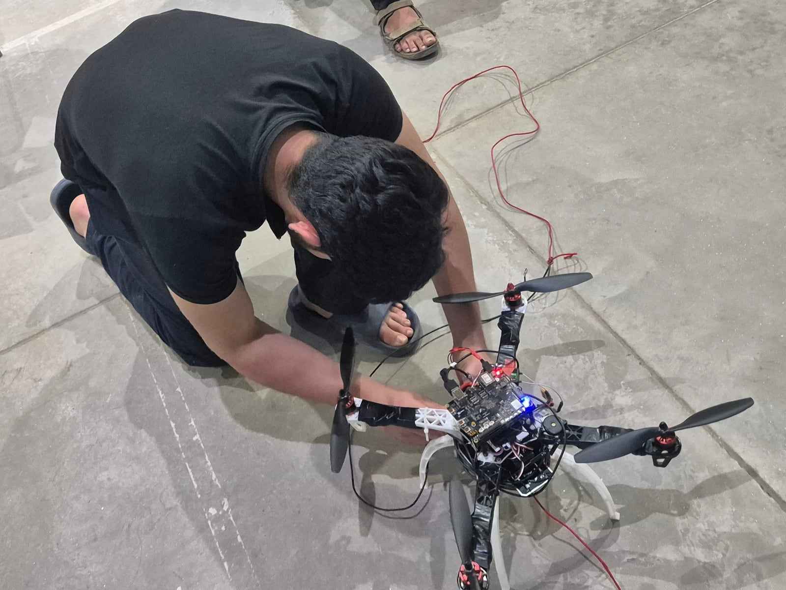 Vedant kneeling on the concrete floor of a large tent hall, working on the ANAV quadcopter before a test session.