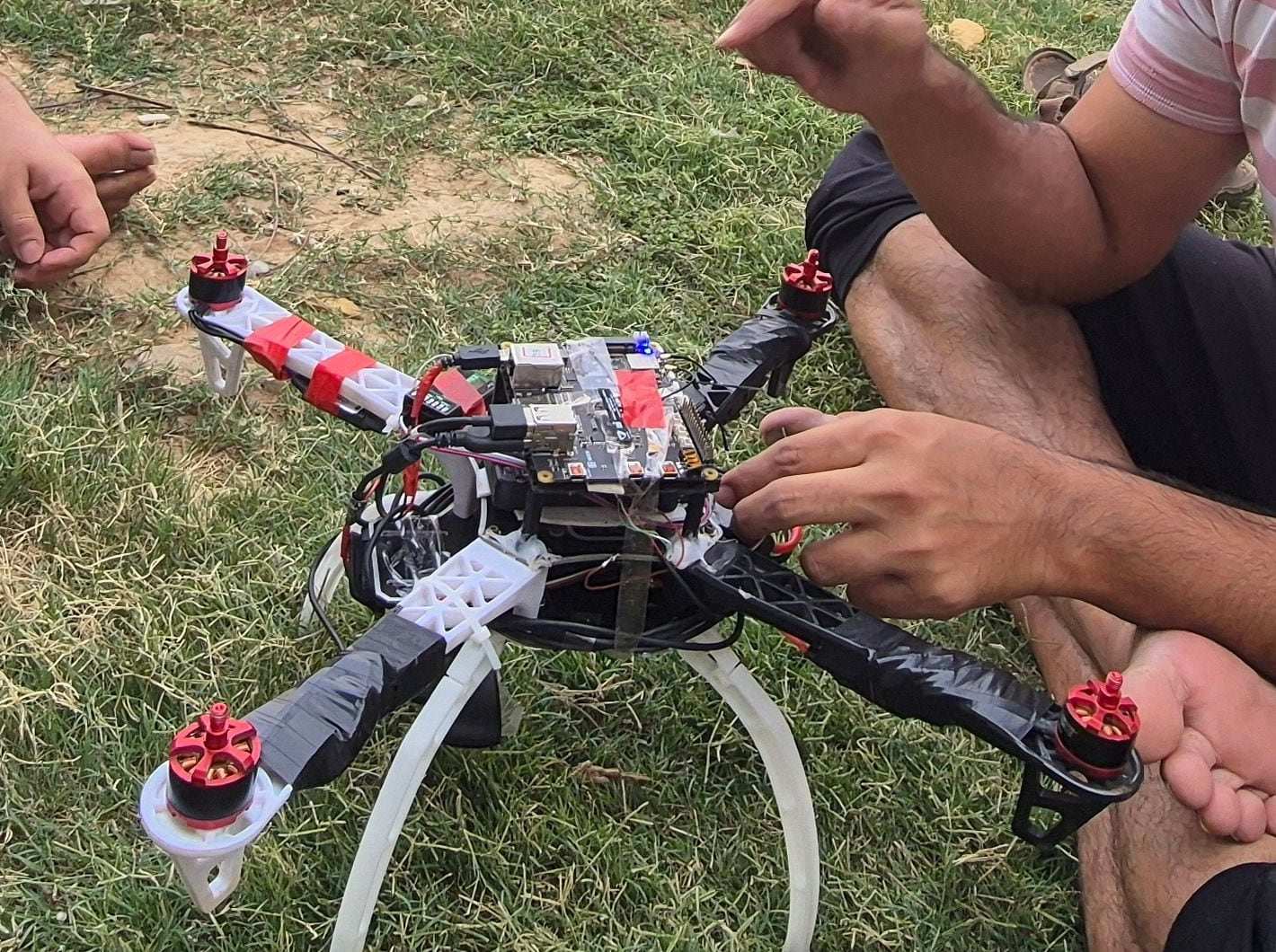 The ANAV quadcopter on grass outdoors, with a teammate's hands adjusting the wiring.