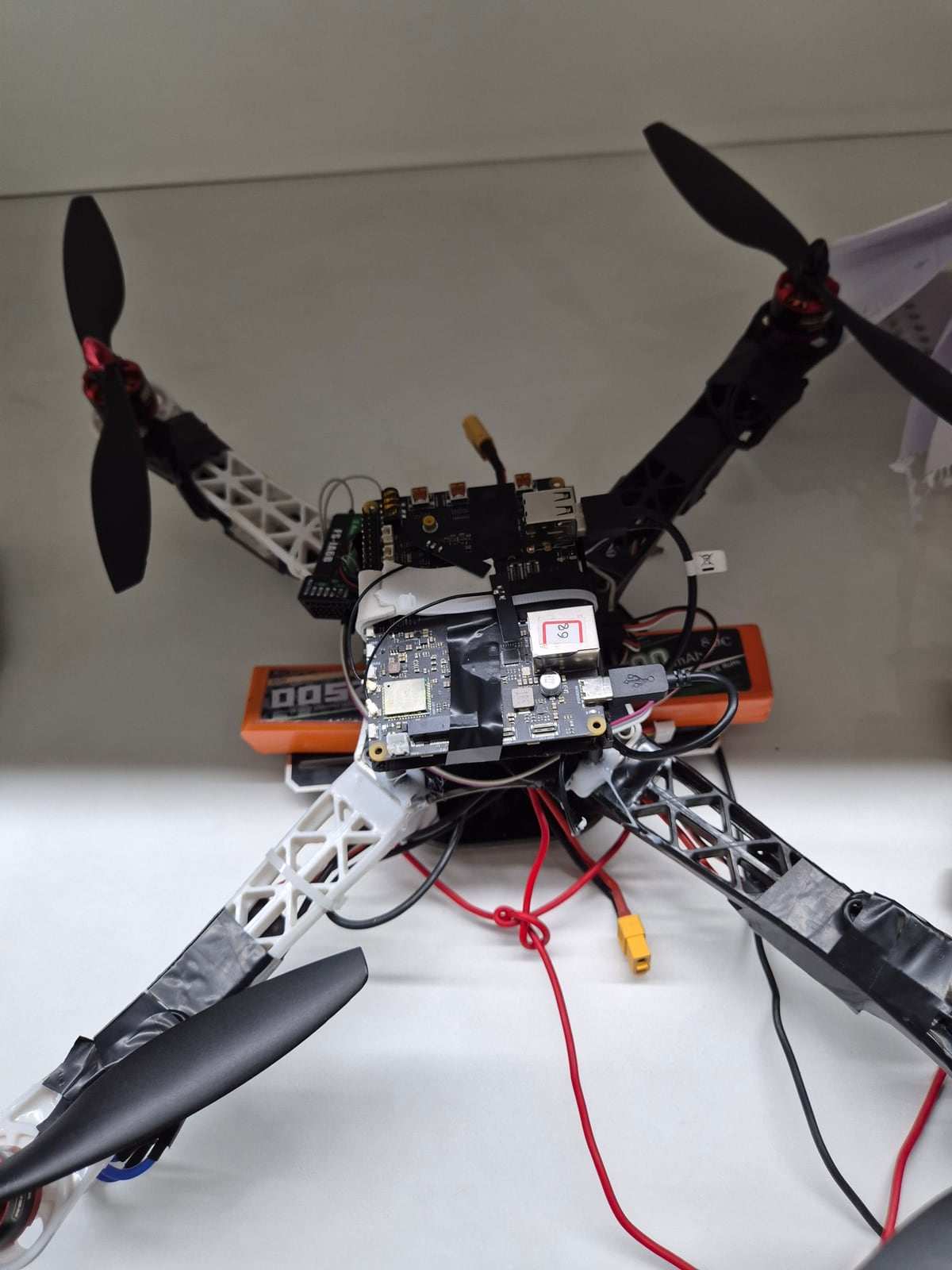 The ANAV quadcopter on a desk, front view: printed white and black arms, a stacked companion computer on top, an FS-iA6B receiver, and an orange LiPo pack underneath.