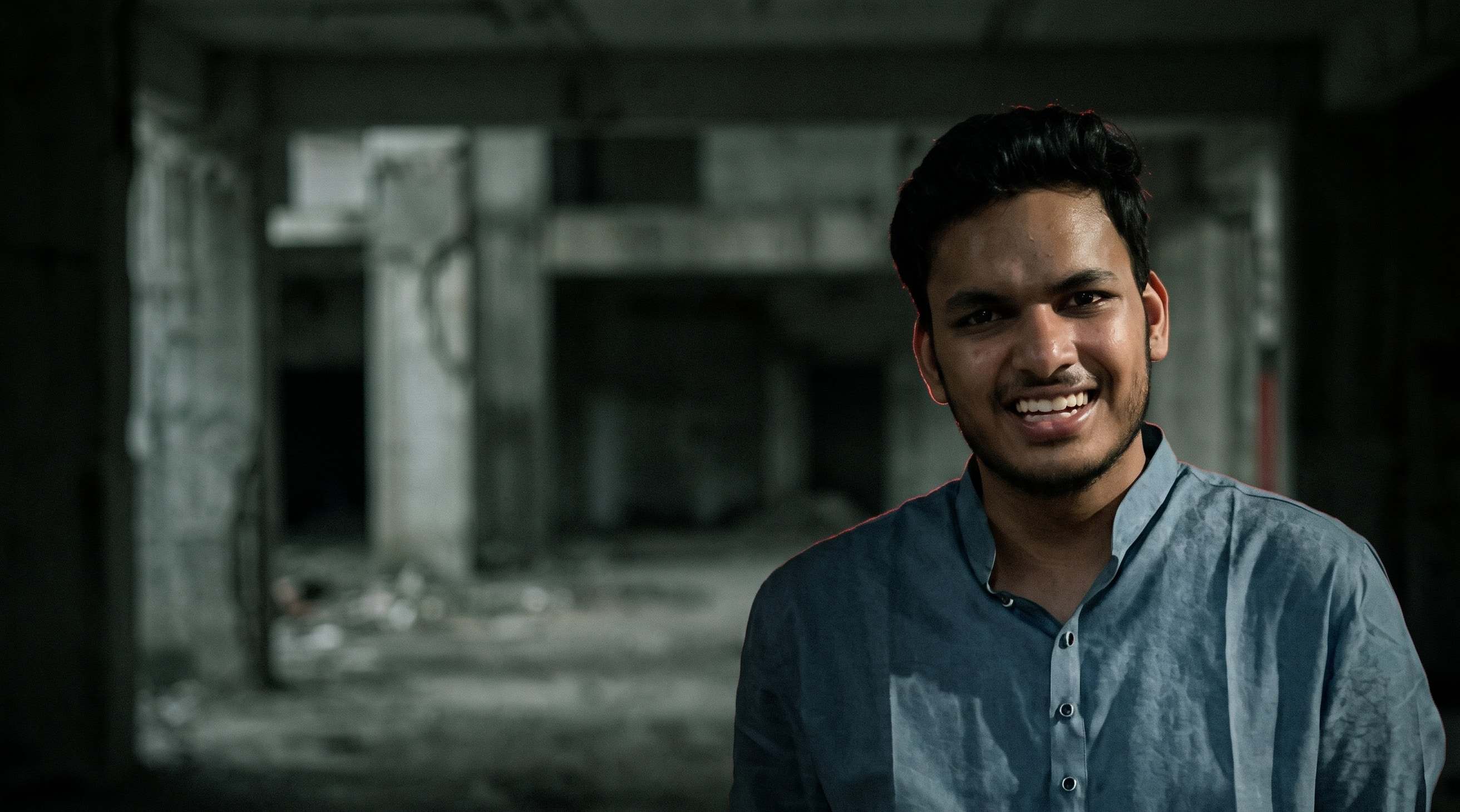 Vedant Somani, smiling, in a blue shirt, standing in a dark, empty concrete hall. Background and lighting AI-edited.